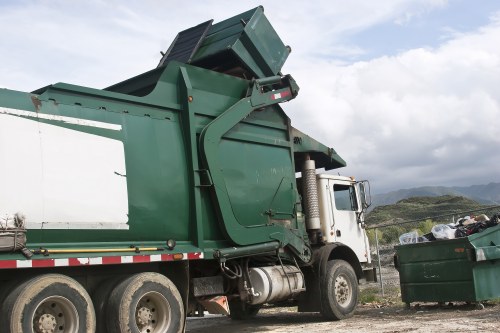 A large industrial waste collection truck with a green and white body is positioned on a gravel surface, with the rear compactor lifting a metal dumpster that is partially open, revealing dark waste materials inside. The truck's cabin is white, and the vehicle has multiple wheels visible beneath the body, including dual rear wheels. The backdrop features a partly cloudy sky and distant rolling hills, suggesting a rural or suburban environment. To the right of the truck, there is a green wheeled bin filled with waste, positioned near the vehicle for collection or disposal. The scene is typical of rubbish removal operations in a local area, with the truck clearly engaged in waste loading activities. The overall atmosphere emphasizes professional rubbish collection, consistent with services offered by companies like Garden Clearance Golders Green, in the context of garden or general rubbish clearance in the London Borough of Barnet area. The elements visually communicate the process of waste disposal in an outdoor setting with natural light illuminating the scene.