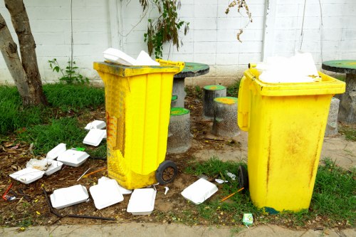 Two yellow wheeled rubbish bins standing on a patch of grass and pavement near a white wooden fence, with both bins open and contaminated with discarded food packaging, including white foam containers spilling onto the ground. The area shows signs of recent waste removal activity, with scattered empty containers and debris surrounding the bins, and a few small trees and plants visible in the background. The scene reflects an outdoor urban or suburban environment, possibly associated with garden or waste clearance services. Garden Clearance Golders Green offers rubbish collection and disposal, as illustrated by the scene of overflowing waste containers and scattered litter in the vicinity of the bins.