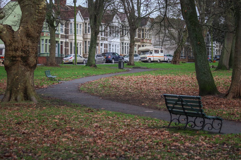 A quiet outdoor scene in a park shows a wooden bench with a weathered, light-colored finish positioned on a concrete pad beside a black metal litter bin labeled 'LITTER'. The bench is situated near a 