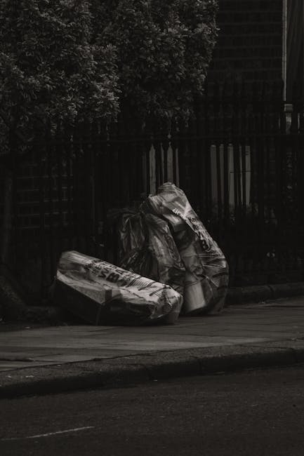 A collection of overflowing rubbish bins and discarded waste materials situated on a paved urban sidewalk next to a parking area. The waste includes various black, red, and grey bins filled with mixed