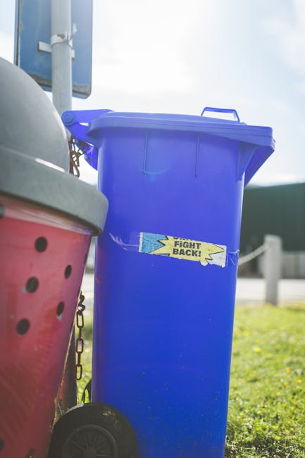 A woman stands on a residential sidewalk next to a row of six large black wheelie bins with flat lids, positioned against the front brick wall of a terraced house. The bins have white chalk markings o